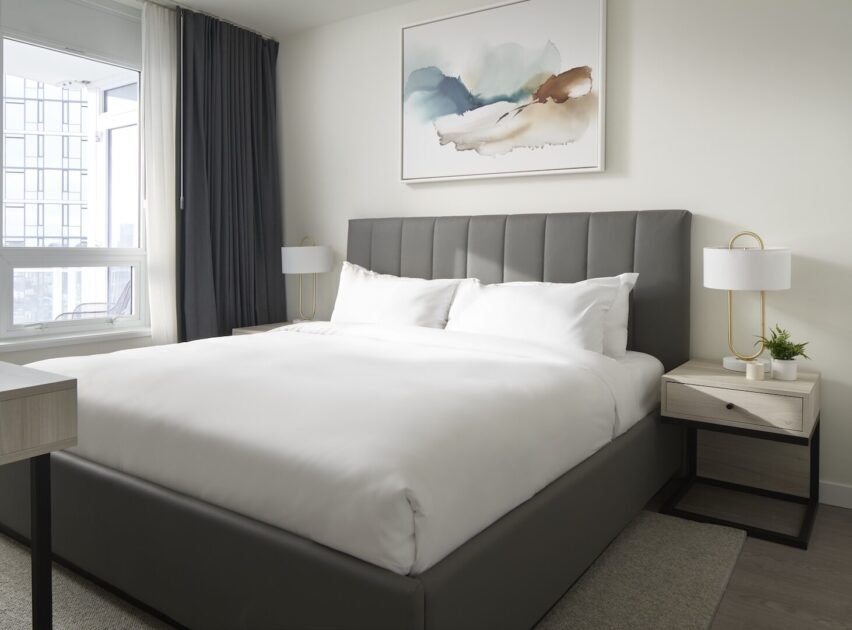 Modern bedroom with a large gray upholstered bed, white bedding, two light wood nightstands with lamps, a window with dark curtains, and abstract art on the wall above the bed.