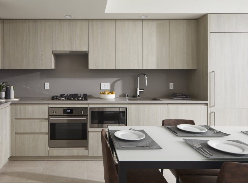 Modern kitchen with light wood cabinets, stainless steel appliances, a built-in oven, gas cooktop, and sink. A dining table with four place settings is in the foreground. The space looks clean and minimalistic.
