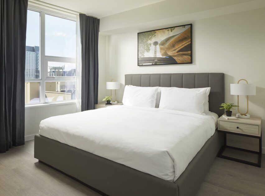 Modern bedroom with a large bed, white bedding, padded grey headboard, two bedside tables with lamps and small plants, large window with city view, dark curtains, and a framed photo on the wall above the bed.