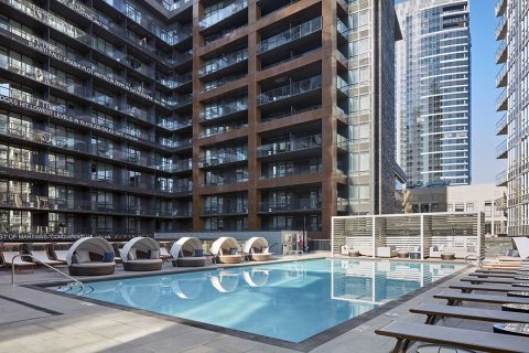 Outdoor swimming pool surrounded by modern high-rise buildings, with lounge chairs and shaded cabanas along the poolside on a sunny day.