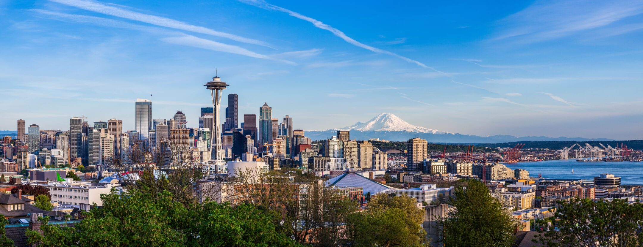 Panoramic view of Seattle skyline with the Space Needle, tall buildings, and Mount Rainier in the background under a blue sky with wispy clouds. Green trees and water are visible in the foreground and to the right.