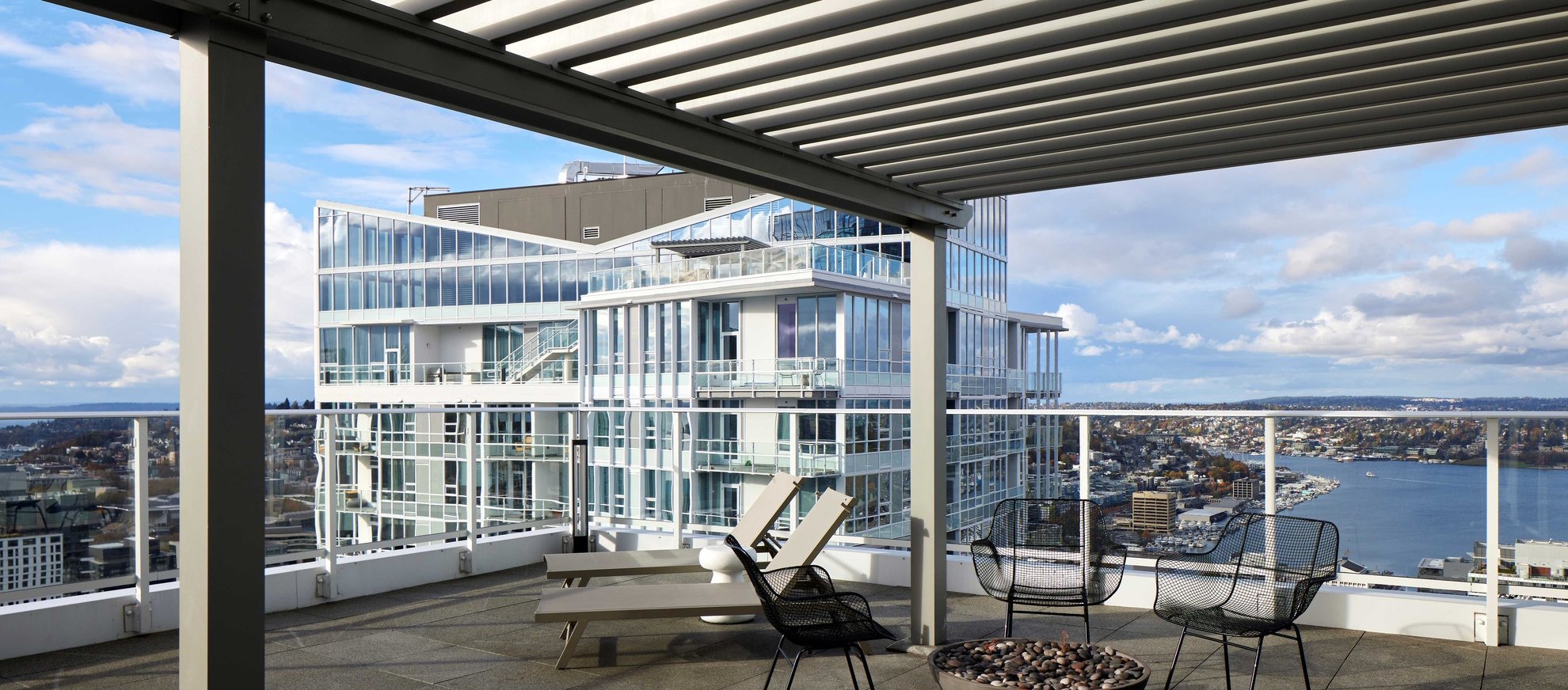 A rooftop patio with modern lounge chairs, a fire pit, and a slatted pergola overlooks a cityscape with a glass building and a body of water under a partly cloudy sky.
