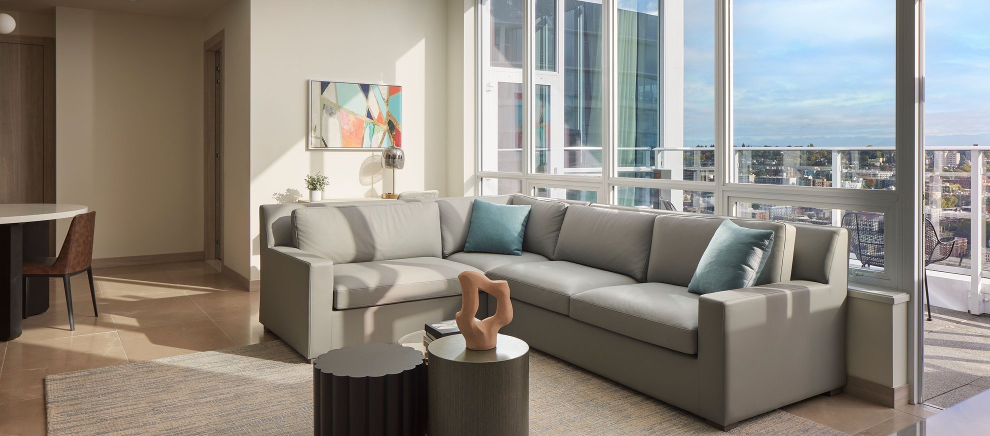 Modern living room with a large light gray sectional sofa, blue and gray pillows, abstract wall art, a sculptural decor piece on a round coffee table, and floor-to-ceiling windows overlooking a cityscape and blue sky.