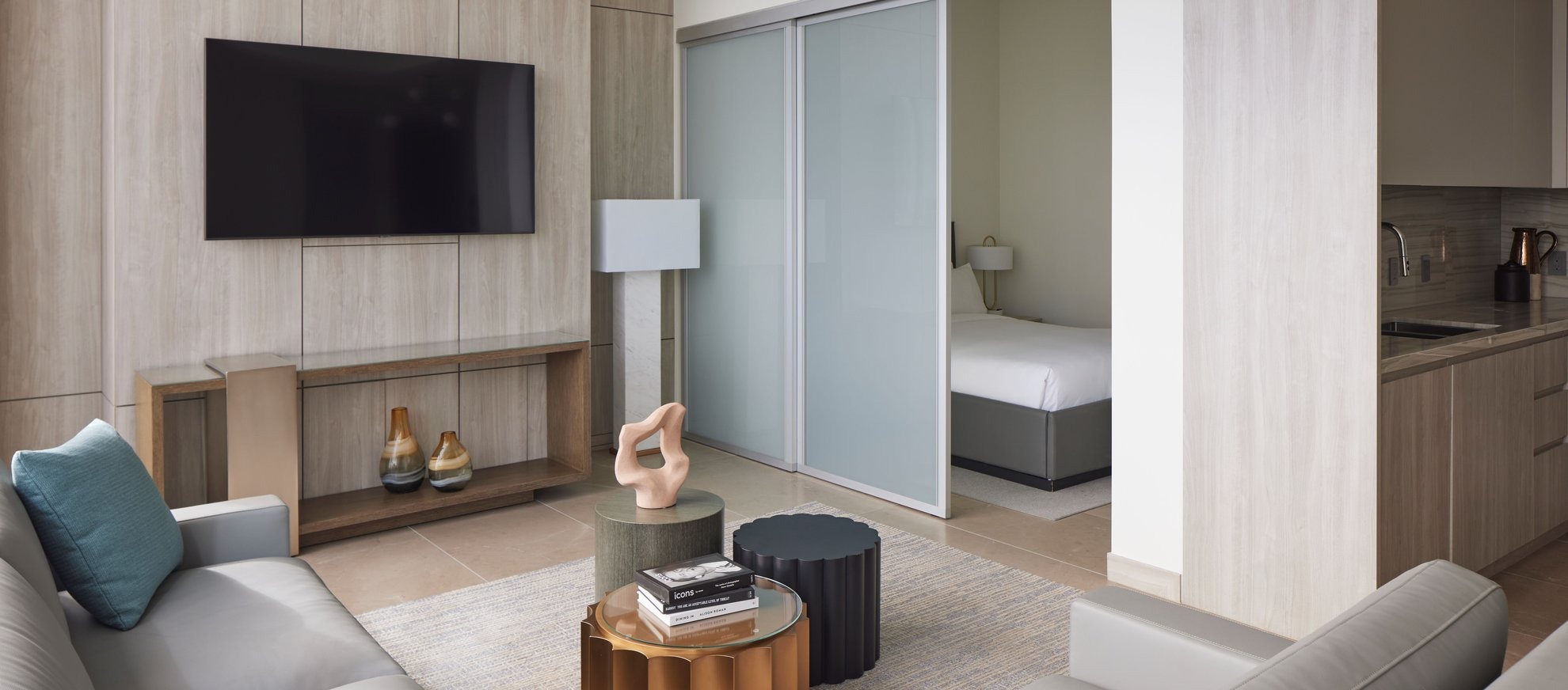 Modern hotel suite with neutral tones, featuring a living area with a couch, armchair, TV, decorative objects, and a coffee table; a bedroom with a bed visible through sliding glass doors, and a kitchenette on the right.