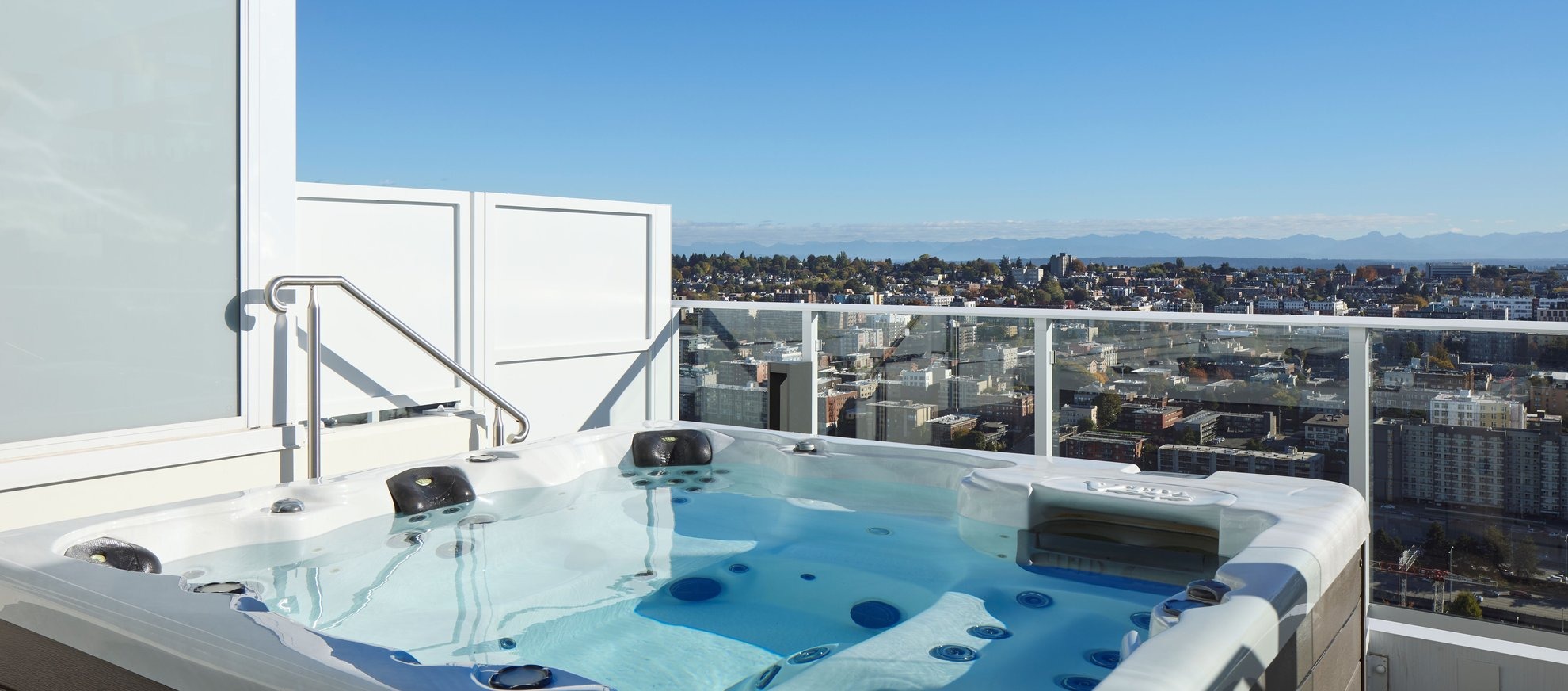 A rooftop hot tub with clear water overlooks a cityscape and distant mountains under a bright blue sky, surrounded by glass railings and a privacy panel.
