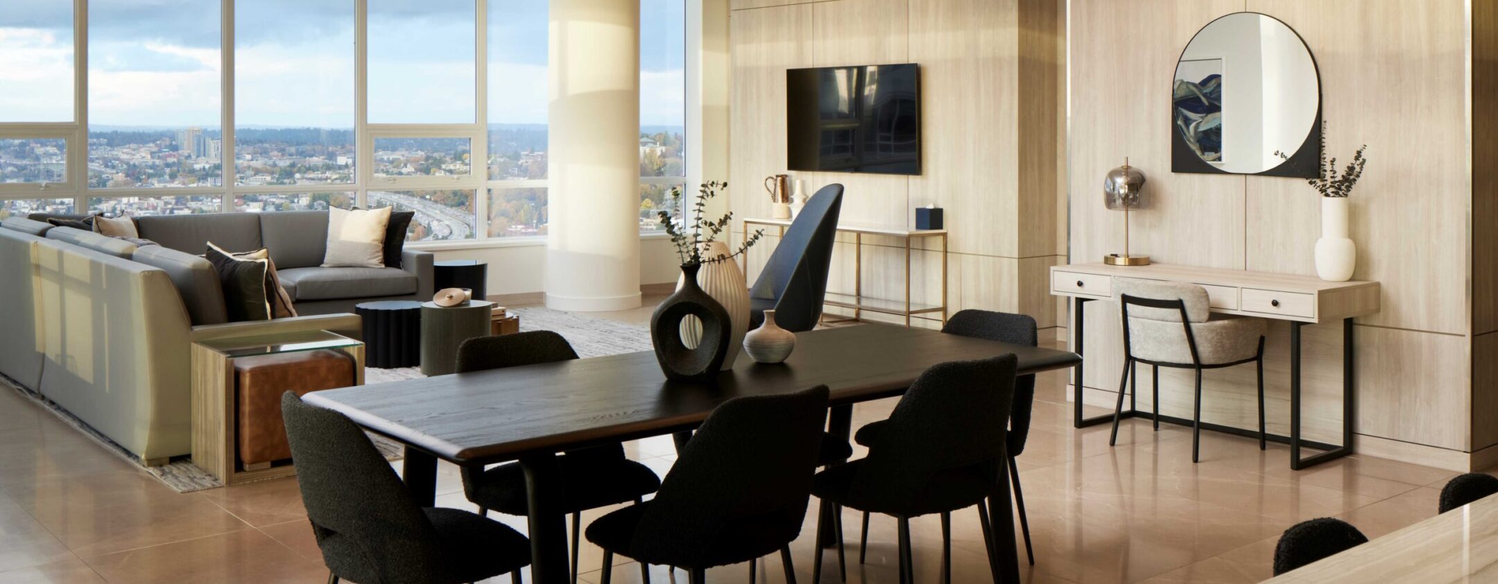 Modern, open-concept living and dining area with large windows, city views, beige walls, a sectional sofa, dark dining set, contemporary light fixture, wall-mounted TV, and a desk with a round mirror and decorative items.