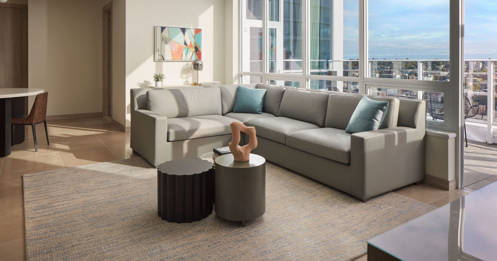 A modern living room with a large gray sectional sofa, two round coffee tables, abstract wall art, and floor-to-ceiling windows offering a city view. A neutral rug and blue accent pillows complete the space.