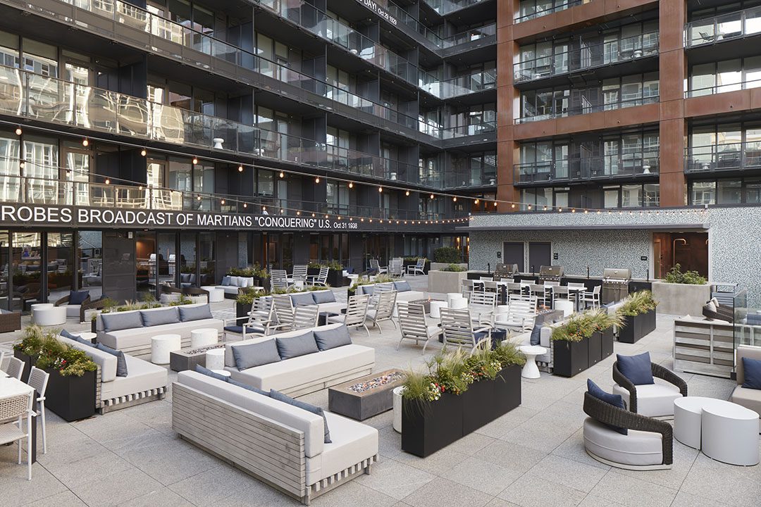 Modern outdoor patio of a hotel with gray sofas, tables, and chairs arranged for seating. Large planters with greenery line the area. Glass balconies overlook the space, and string lights hang above.