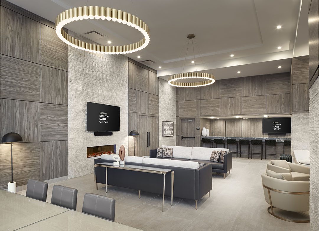 A modern lounge with gray wood paneling, a wall-mounted TV, black couches, a light stone accent wall with a fireplace, a kitchen area with barstools, and two large circular chandeliers.