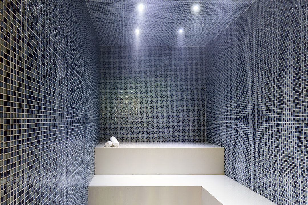 A modern steam room with blue mosaic tile walls and ceiling, white benches, and rolled white towels under soft overhead lighting.