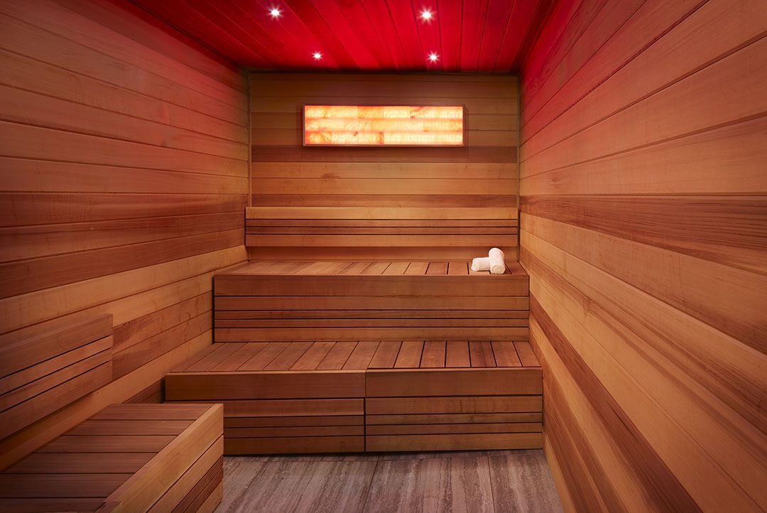 A modern wooden sauna room with tiered seating, red ceiling lights, and a small window. Rolled white towels are placed on the top bench, and the floor is made of light-colored wood.