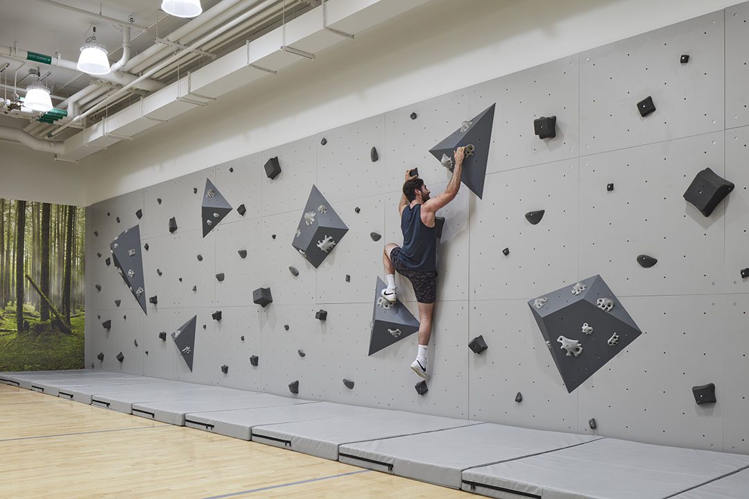 A person climbs an indoor bouldering wall with geometric black holds and volumes, on a padded gym floor. The wall is light gray, and there is a forest mural on the side wall.