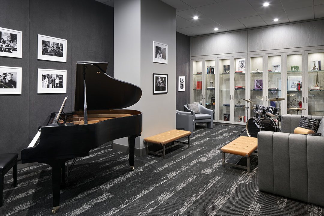 A modern music room with a grand piano, drum set, gray chairs, sofas, and glass display cases. Black-and-white photos decorate dark walls, and spotlights brighten the stylish, carpeted space.