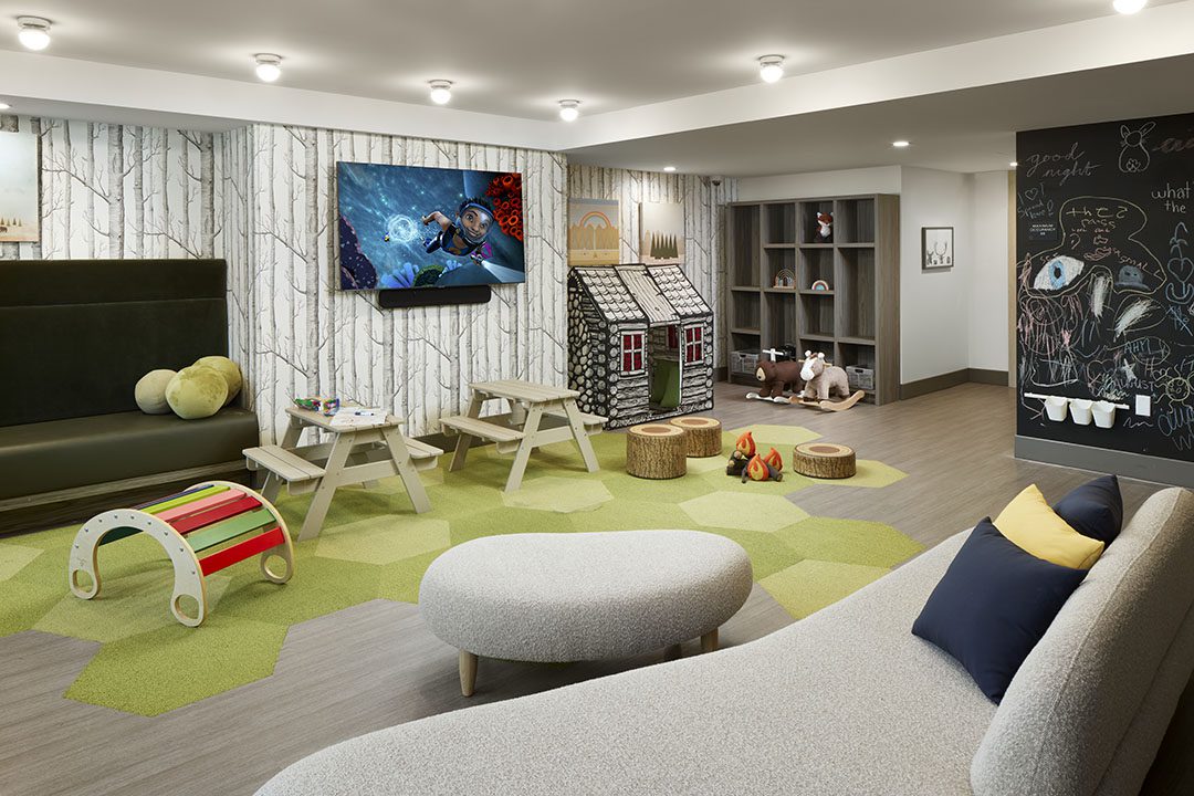 Modern children’s playroom with green and tan flooring, small tables and benches, a TV on the wall, a playhouse, a reading nook, toys, bookshelves, and a chalkboard wall decorated with drawings.