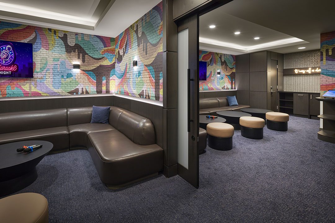 Modern karaoke lounge with brown leather sofas, round ottomans, colorful graffiti-style wall art, dark tables, a glass partition, and neon signs on the walls, creating a lively and inviting atmosphere.