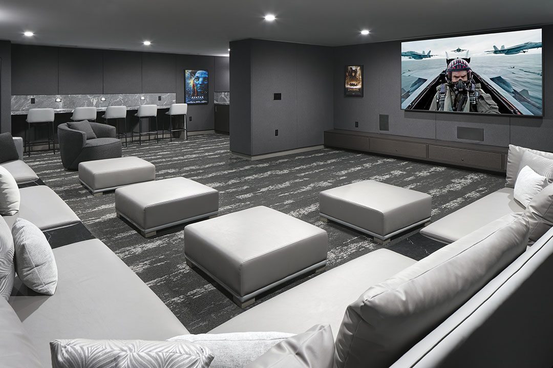 Modern home theater room with gray sofas, ottomans, and a large screen displaying a fighter jet scene. Movie posters decorate the walls, and a bar area with stools is seen in the back. The room is stylish and well-lit.