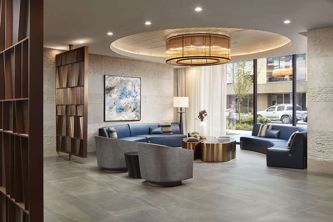 Modern hotel lobby with a large circular ceiling light, blue and gray seating, a round coffee table, abstract wall art, and floor-to-ceiling windows letting in natural light, with a view of cars outside.