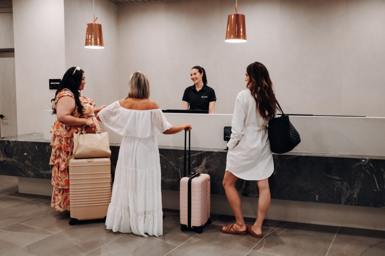 Three women with suitcases stand at a hotel reception desk, speaking to a smiling receptionist behind the counter. The lobby is modern with neutral tones and pendant lights overhead.