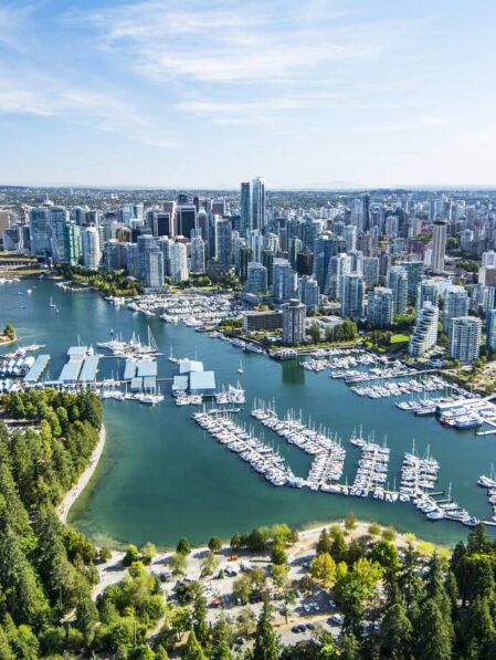 Aerial view of Vancouver with high-rise buildings, marinas full of boats, tree-lined Stanley Park in the foreground, and blue water under a clear sky.