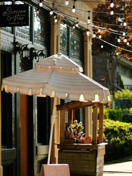 A sunlit sidewalk scene featuring a small wooden flower stand with a striped umbrella, string lights overhead, and a sign reading “Blossom & Wine.” The stand displays bouquets and is set in front of a charming storefront.