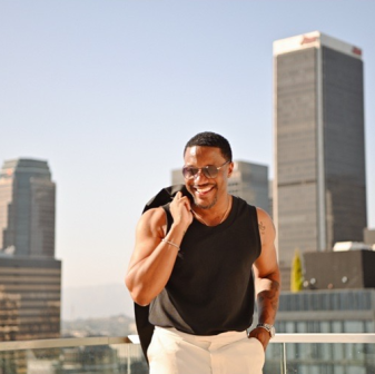 A smiling man in sunglasses and a sleeveless black shirt holds a jacket over his shoulder, standing on a rooftop with tall city buildings in the background.