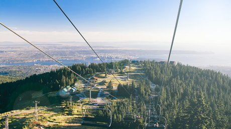 A scenic view from a ski lift descending over a forested mountain, overlooking a city, water, and distant land under a clear blue sky.