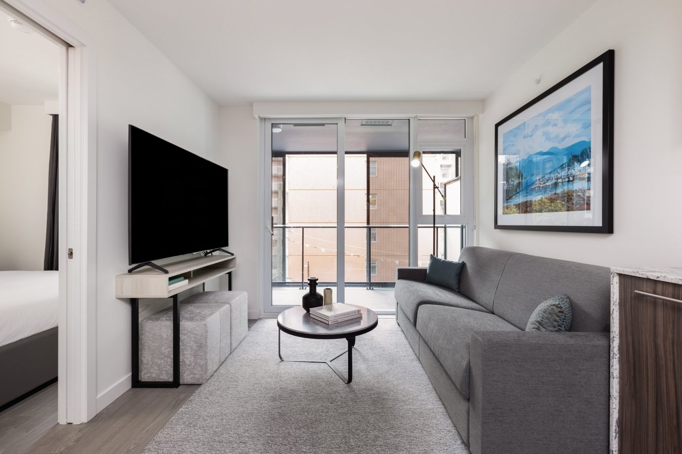 Modern living room with a gray sofa, coffee table, large flat-screen TV, and a framed mountain landscape photo on the wall. Floor-to-ceiling windows open to a balcony with city views.