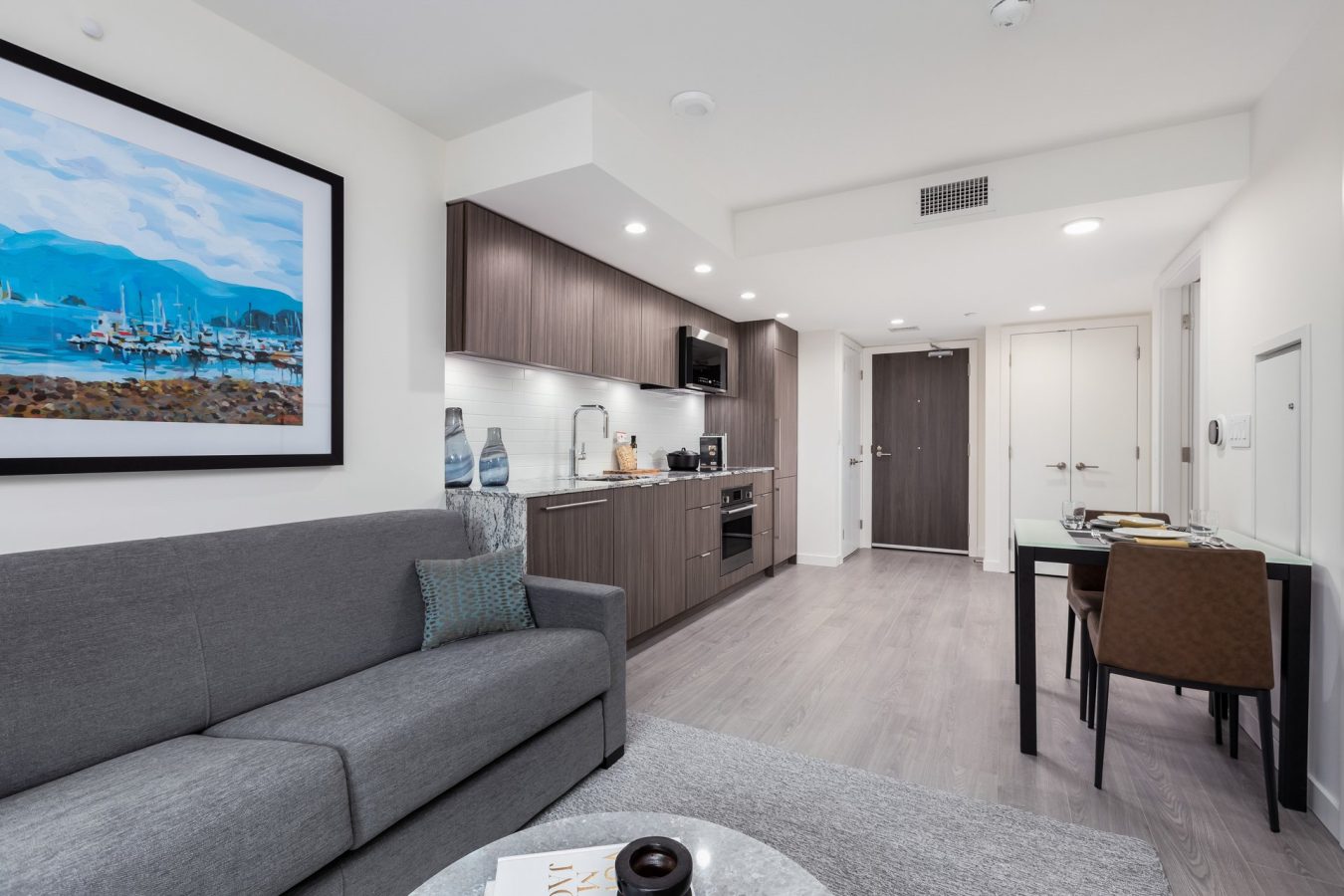 Modern studio apartment interior featuring a gray sofa, wall art, kitchenette with wood cabinets, a dining table with two chairs, and a light wood floor, all in a bright, minimalist setting.