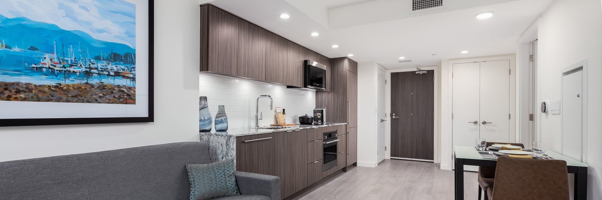 Modern apartment with a compact kitchen, gray cabinets, a dining table set for two, and a sofa. A colorful painting of boats hangs on the wall above the sofa. The space is well-lit with recessed ceiling lights.