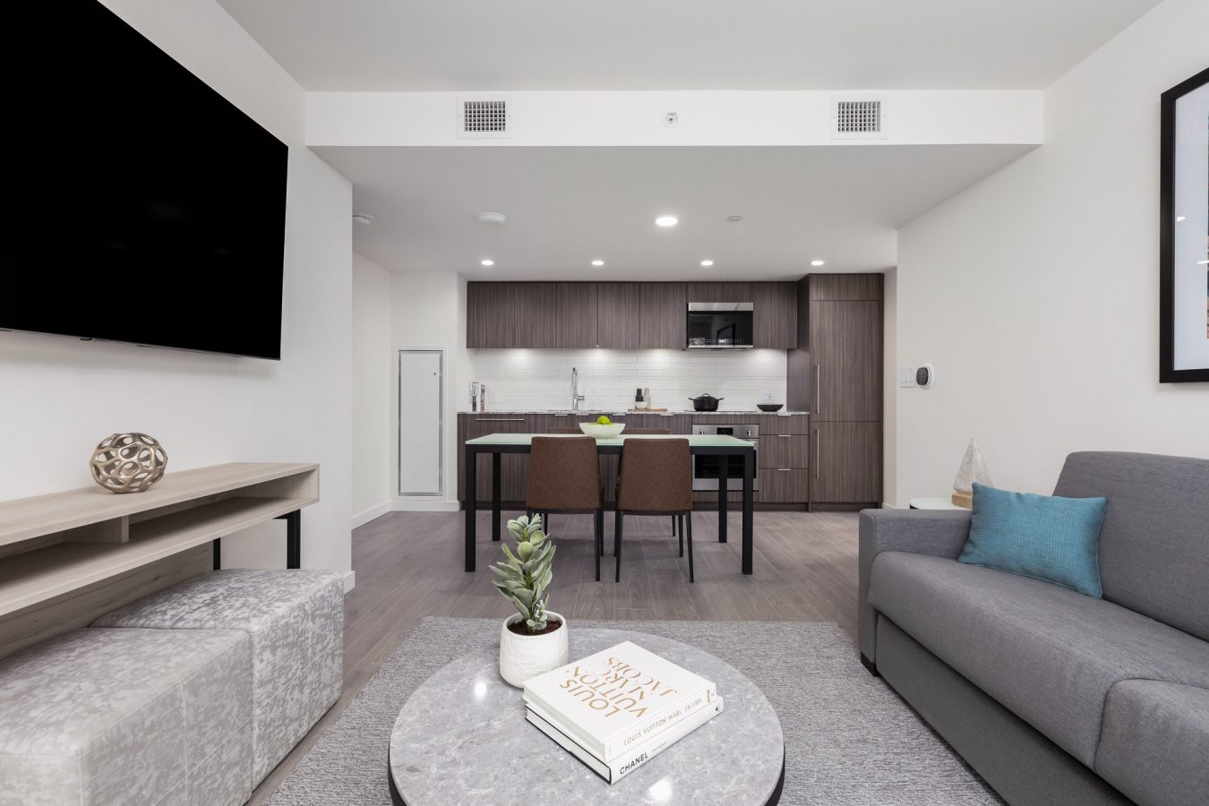 Modern open-plan living room and kitchen with gray sofa, round coffee table, two brown dining chairs, and sleek wood cabinetry. Neutral tones and minimalist decor create a clean, contemporary atmosphere.