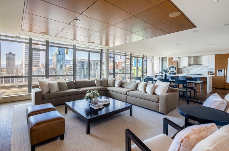 A modern, spacious living room with large windows showing a city view, a beige sectional sofa, two brown ottomans, a black coffee table, and an open kitchen with bar seating in the background.