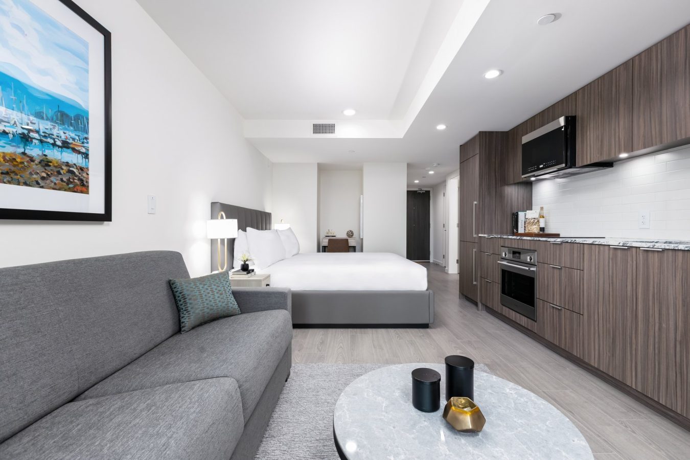 Modern studio apartment with a gray sofa, marble coffee table, kitchenette with wood cabinets, and a bed in the background; wall art and decor create a clean, minimalist look.