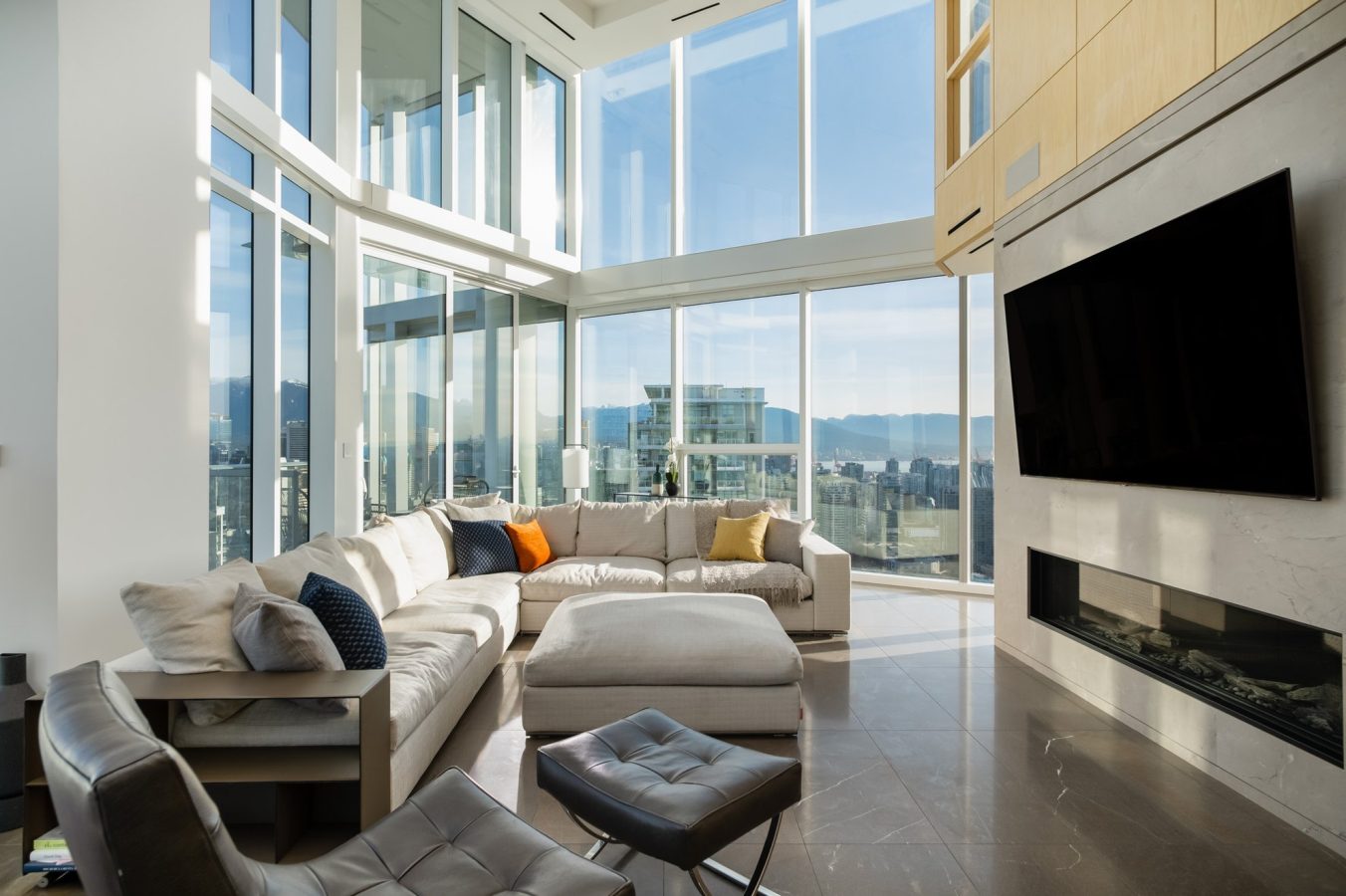 A modern living room with floor-to-ceiling windows, neutral-toned sectional sofa with colorful pillows, large wall-mounted TV, fireplace, and cityscape views in the background. Sunlight fills the bright, open space.