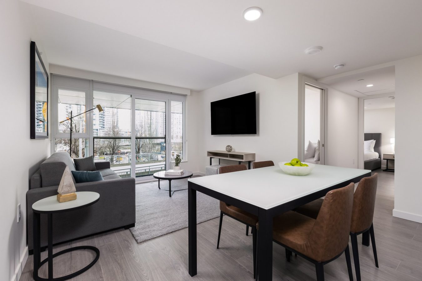 Modern apartment living room with large windows, gray sofa, TV on the wall, and a dining table with four brown chairs. Natural light brightens the space, and a bedroom is visible through an open doorway.