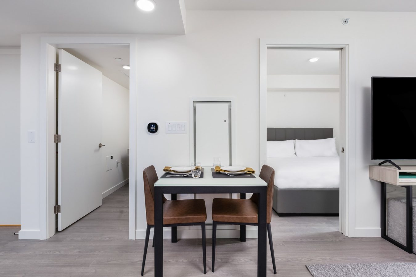 A modern apartment interior with a small dining table set for two, two brown chairs, a bedroom with a bed in the background, and a hallway with white doors. Neutral colors and minimalistic decor.