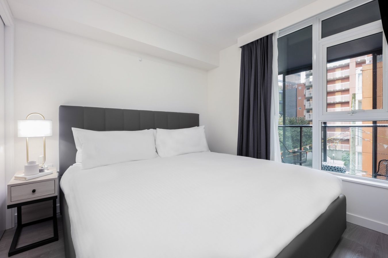 A modern, minimalist bedroom with a large bed featuring white bedding, a dark gray headboard, a small bedside table with a lamp, and floor-to-ceiling windows with dark curtains and a view of buildings outside.
