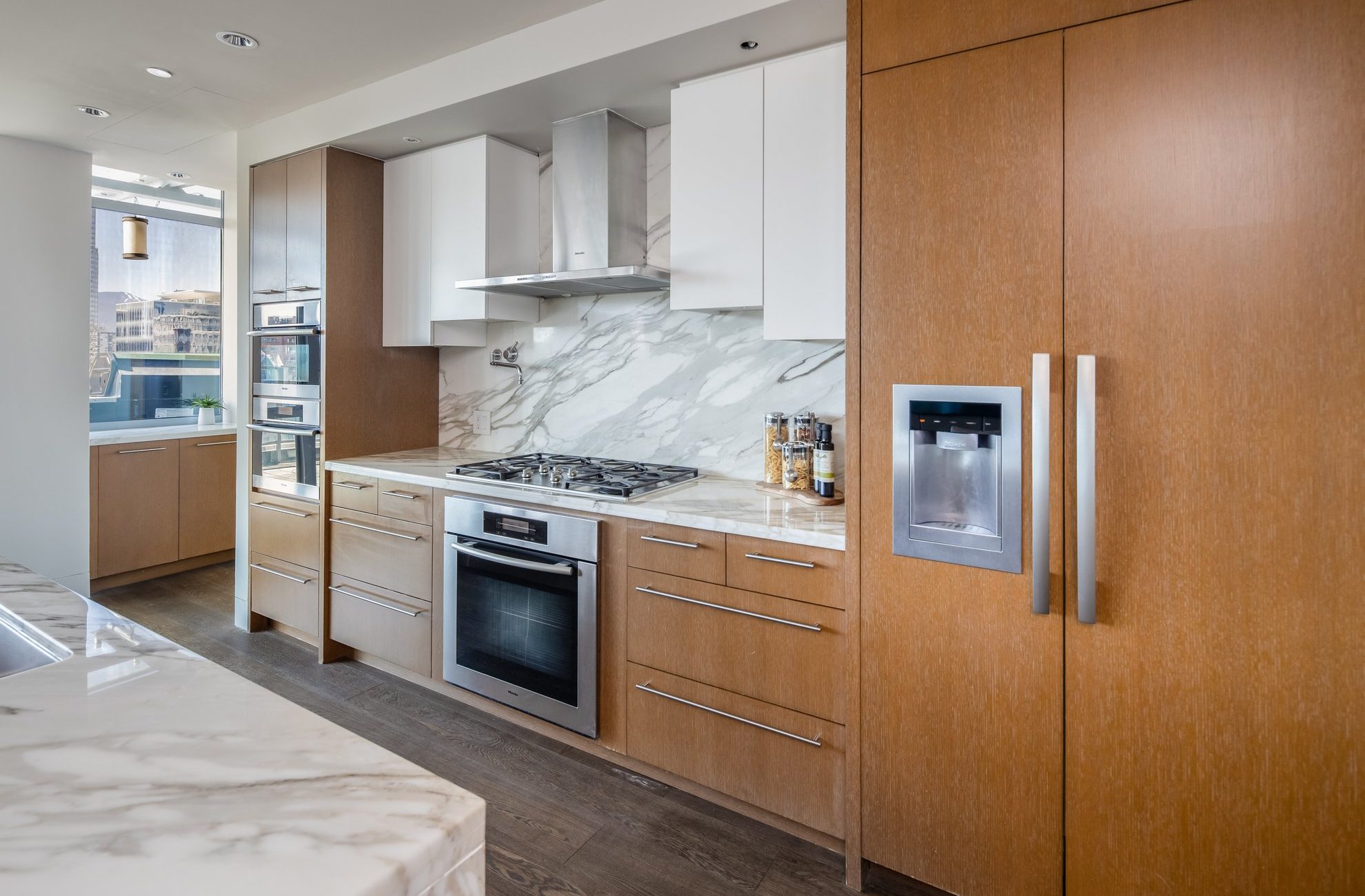 Modern kitchen with wood cabinets, marble countertops and backsplash, stainless steel appliances including a built-in oven, gas cooktop, range hood, and a refrigerator with a water dispenser.