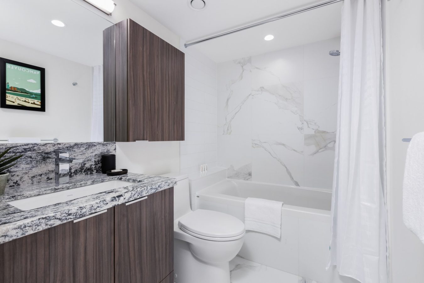 Modern bathroom with a large mirror, woodgrain cabinets, marble countertop and backsplash, a white toilet, a bathtub with marble tile surround, white shower curtain, and a white towel hanging on the wall.