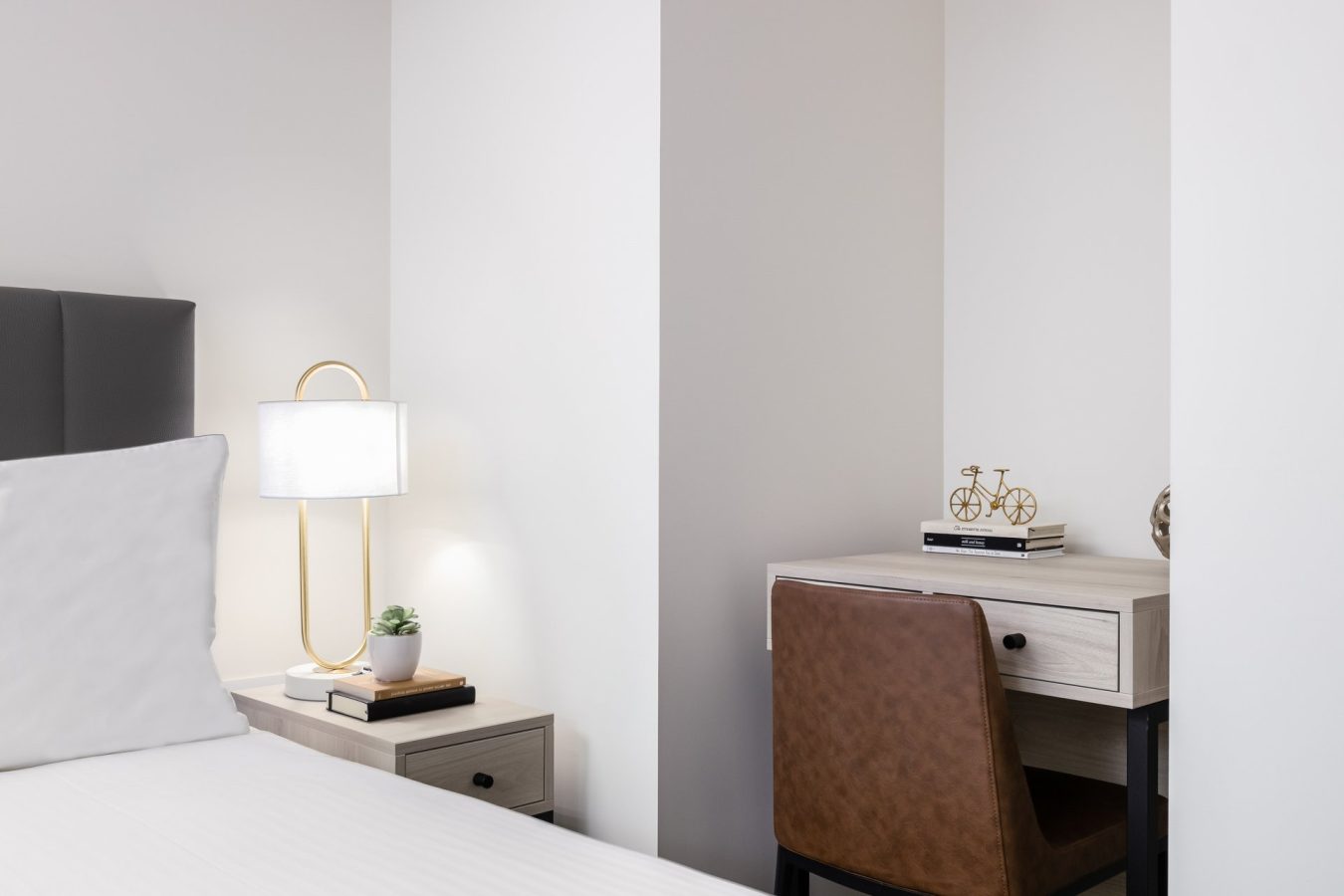 A modern, minimalistic bedroom corner with a bed, a nightstand holding a lamp and books, and a desk with a brown chair, books, a bicycle figurine, and a small plant. The room features white walls and clean lines.