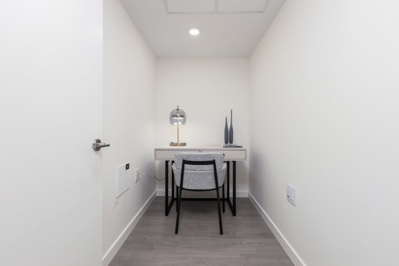 A small, minimalist workspace with a desk, chair, lamp, and two decorative vases, placed against a white wall in a narrow room with light wood flooring.