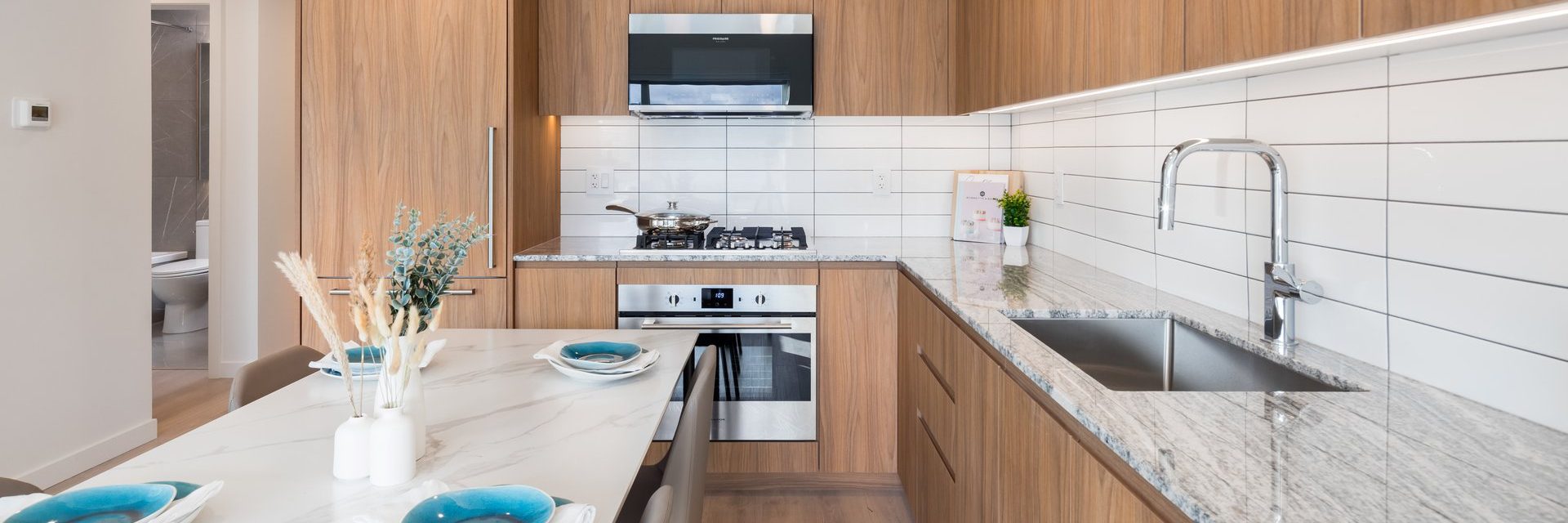 Modern kitchen with wooden cabinets, white tiled backsplash, marble countertops, stainless steel appliances, a sink, and a dining table set with blue and white dishes. A bathroom is visible through an open door.