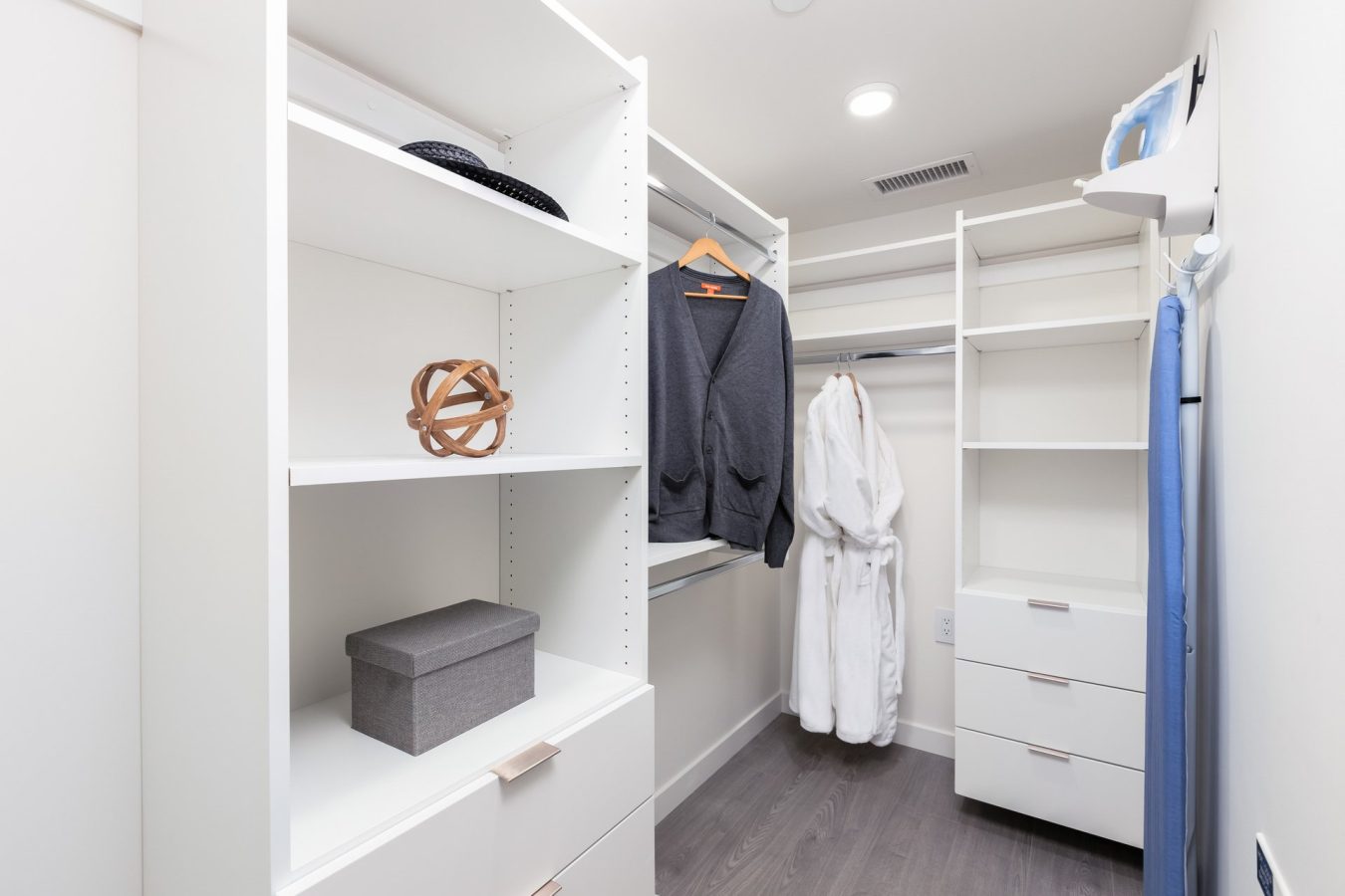 A modern walk-in closet with white shelves, hanging rods, a gray box, a decorative sphere, a gray blazer on a hanger, and a white robe hanging on the back wall. The floor is dark wood.
