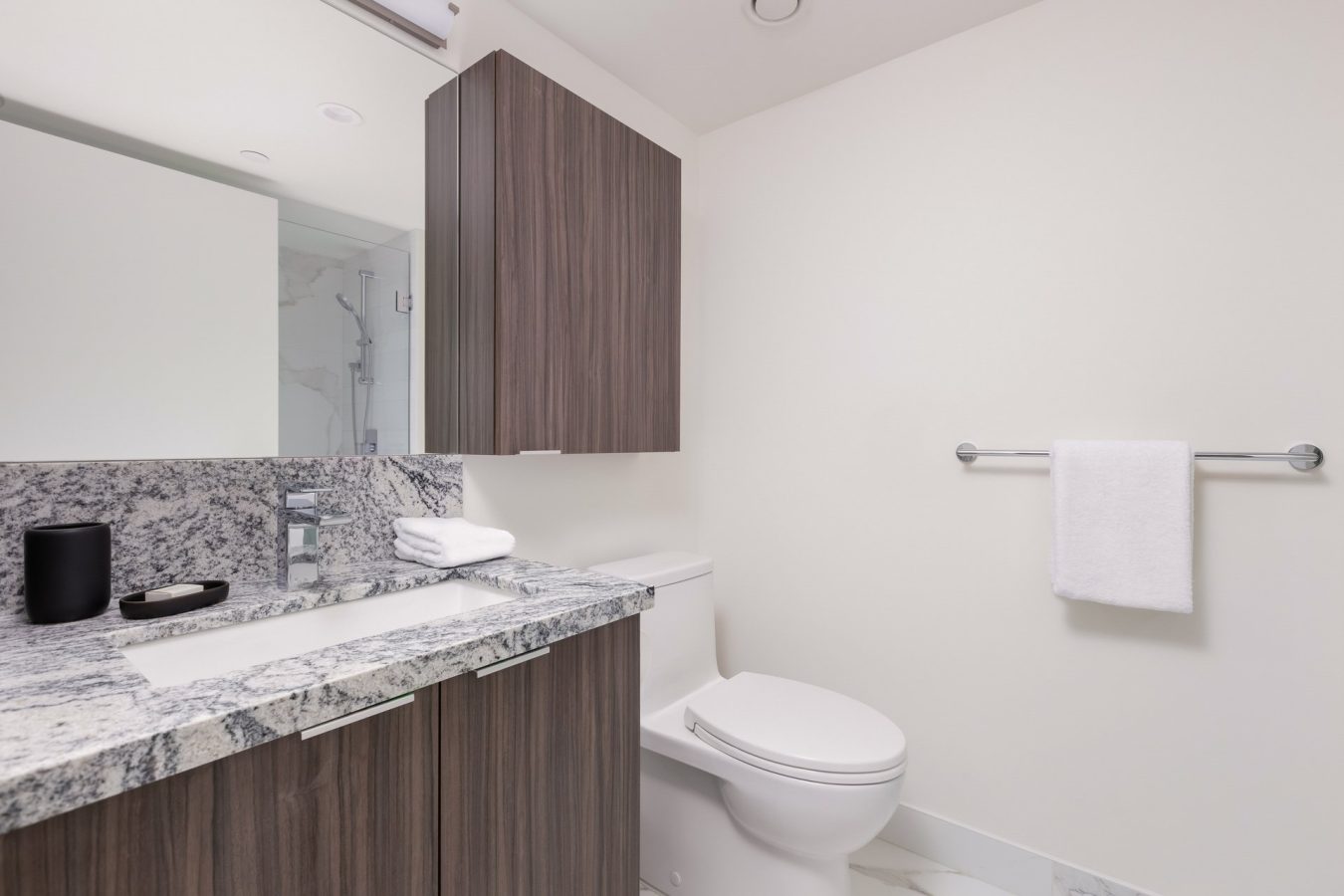Modern bathroom with a granite countertop, sink, and wooden cabinets. A white toilet is next to a towel bar with a folded white towel. The space has white walls and a large mirror above the sink.