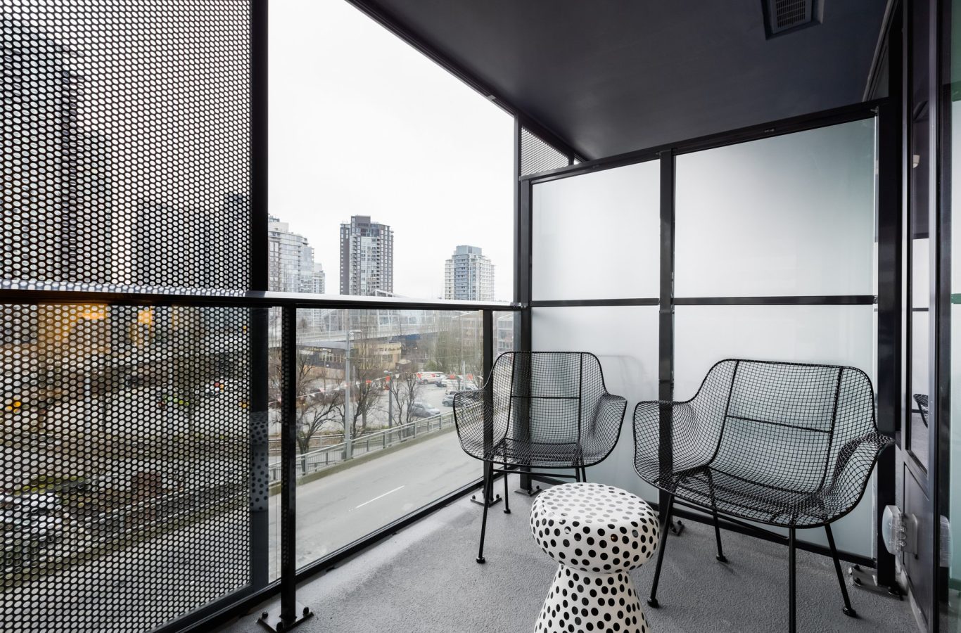A modern apartment balcony with perforated metal railings, two black wire chairs, and a small white table with black polka dots. Urban buildings and a street are visible in the background.