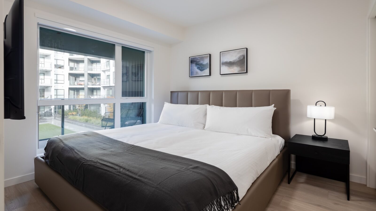 A modern bedroom with a large bed, white bedding, and a dark throw blanket. A nightstand with a lamp sits beside the bed, and two abstract artworks hang above the headboard. A large window shows an apartment building outside.