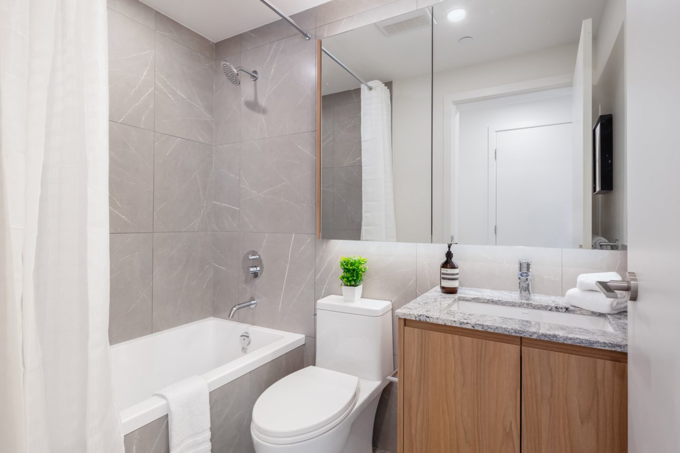 Modern bathroom with gray tiled walls, a white bathtub with a shower, toilet, wooden vanity with a marble countertop, mirror, potted plant, soap dispenser, and folded towels on the sink.