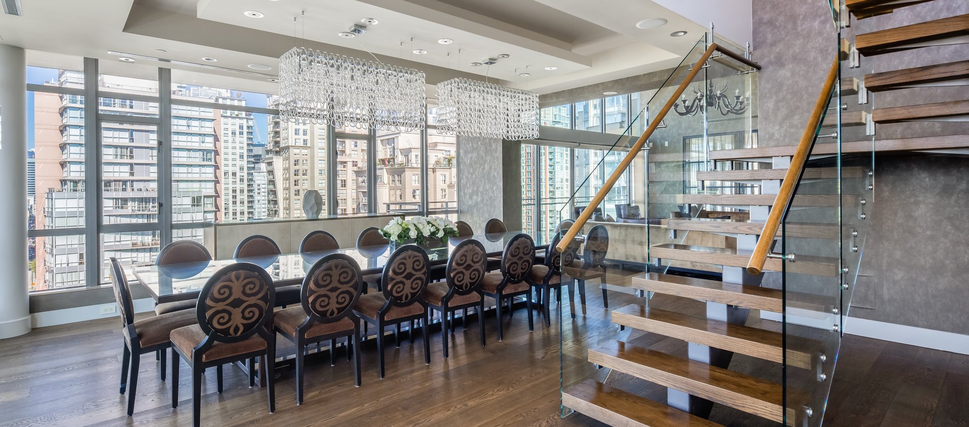Modern dining room with a long table, twelve ornate chairs, two large crystal chandeliers, floor-to-ceiling windows with city views, and a glass staircase with wooden steps.