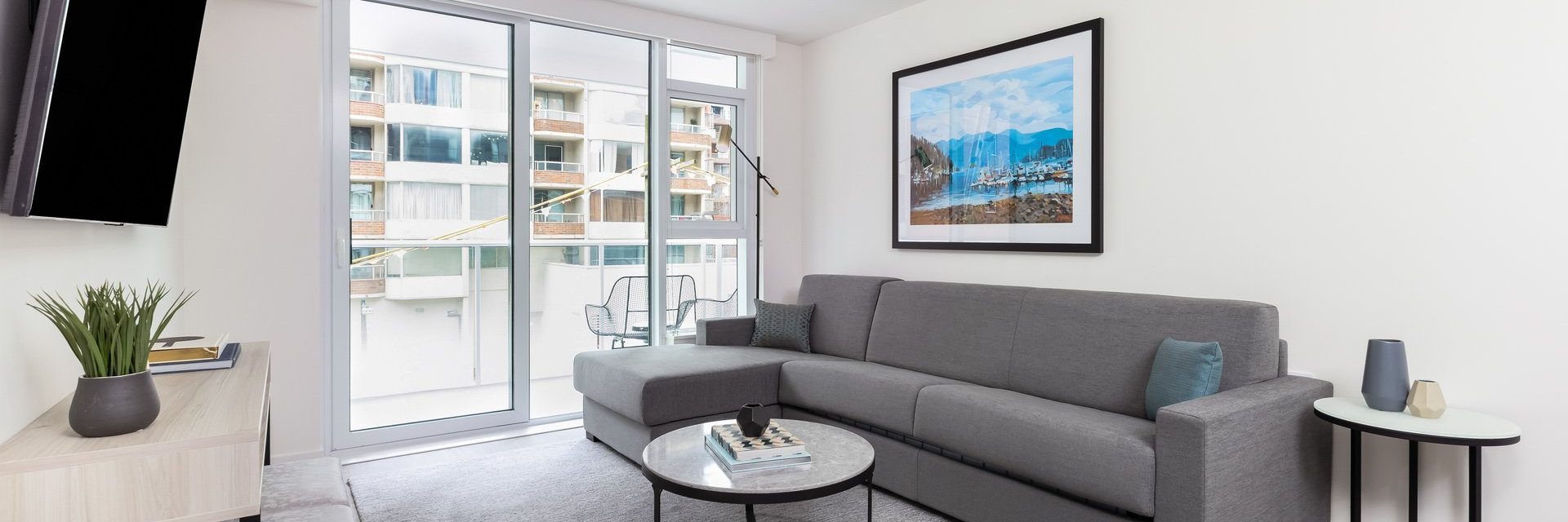 A modern living room with a gray sectional sofa, a round marble coffee table, a TV on a light wood cabinet, and large windows overlooking apartment balconies. A framed landscape photo hangs on a white wall.