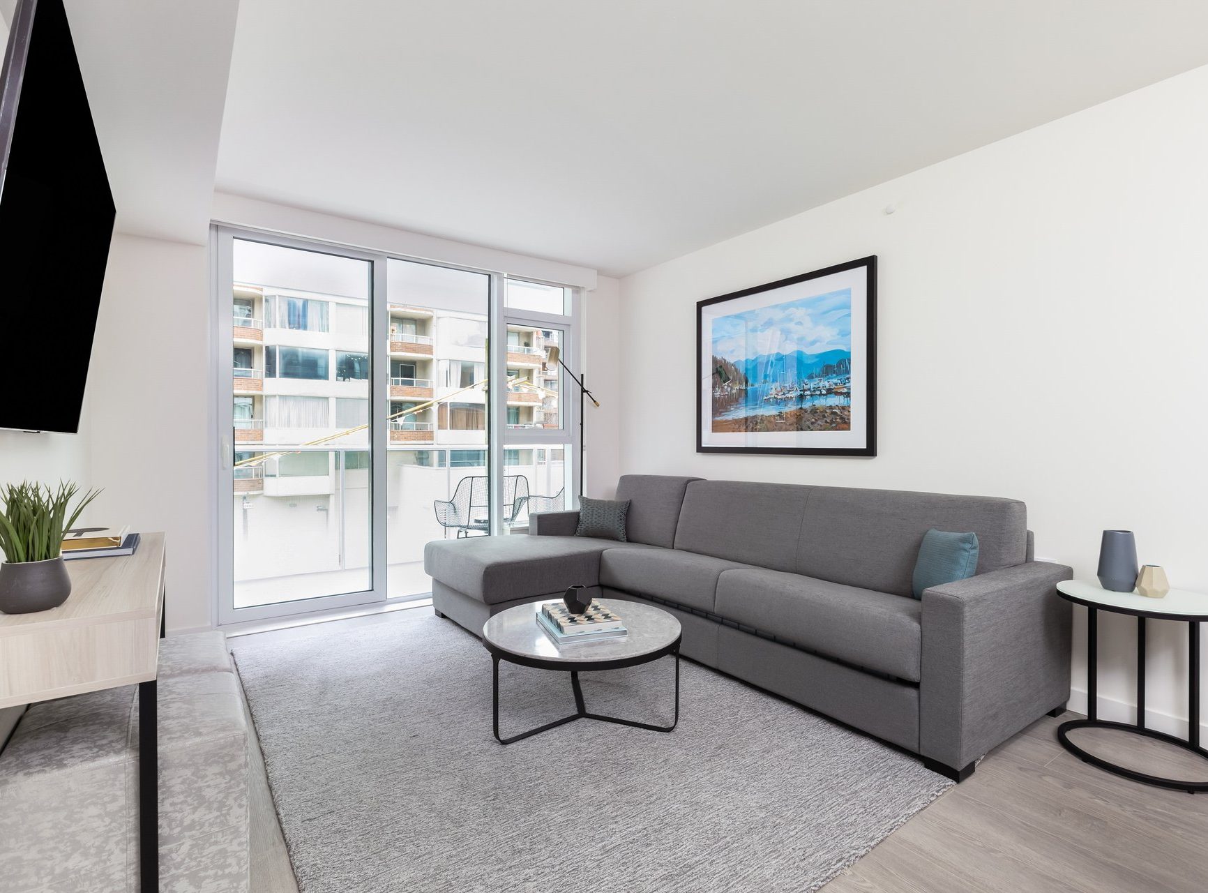 A modern living room with a gray sofa, round coffee table, TV, and a large window that opens to a balcony. Light walls, minimalist decor, and a framed landscape photo create a bright and comfortable space.