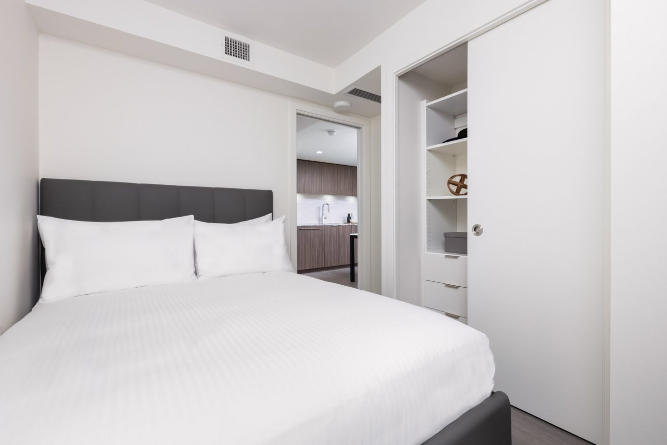 A modern bedroom with a neatly made bed with white linens, an open closet with shelves, and a view into a kitchen area through an open door. The walls are light-colored and the space is minimalistic.
